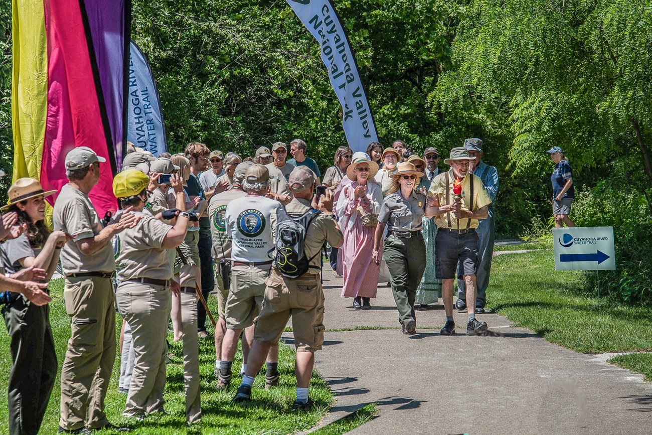 Volunteers and rangers stand and applaud as a ranger and volunteer lead a parade carrying a red-orange facsimile of a torch.