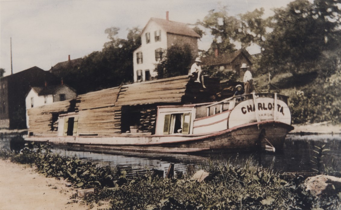 Black and white photo of a canal boat piled high with lumber, floating on the canal; an adult and two children sit near the stern, which reads "Charlotta".