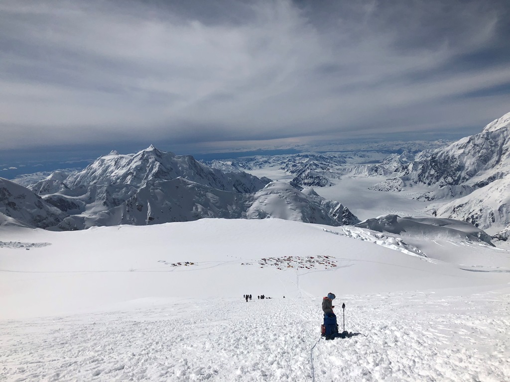 A climber pauses on a hilly slope with climbers and tents down below in the distance