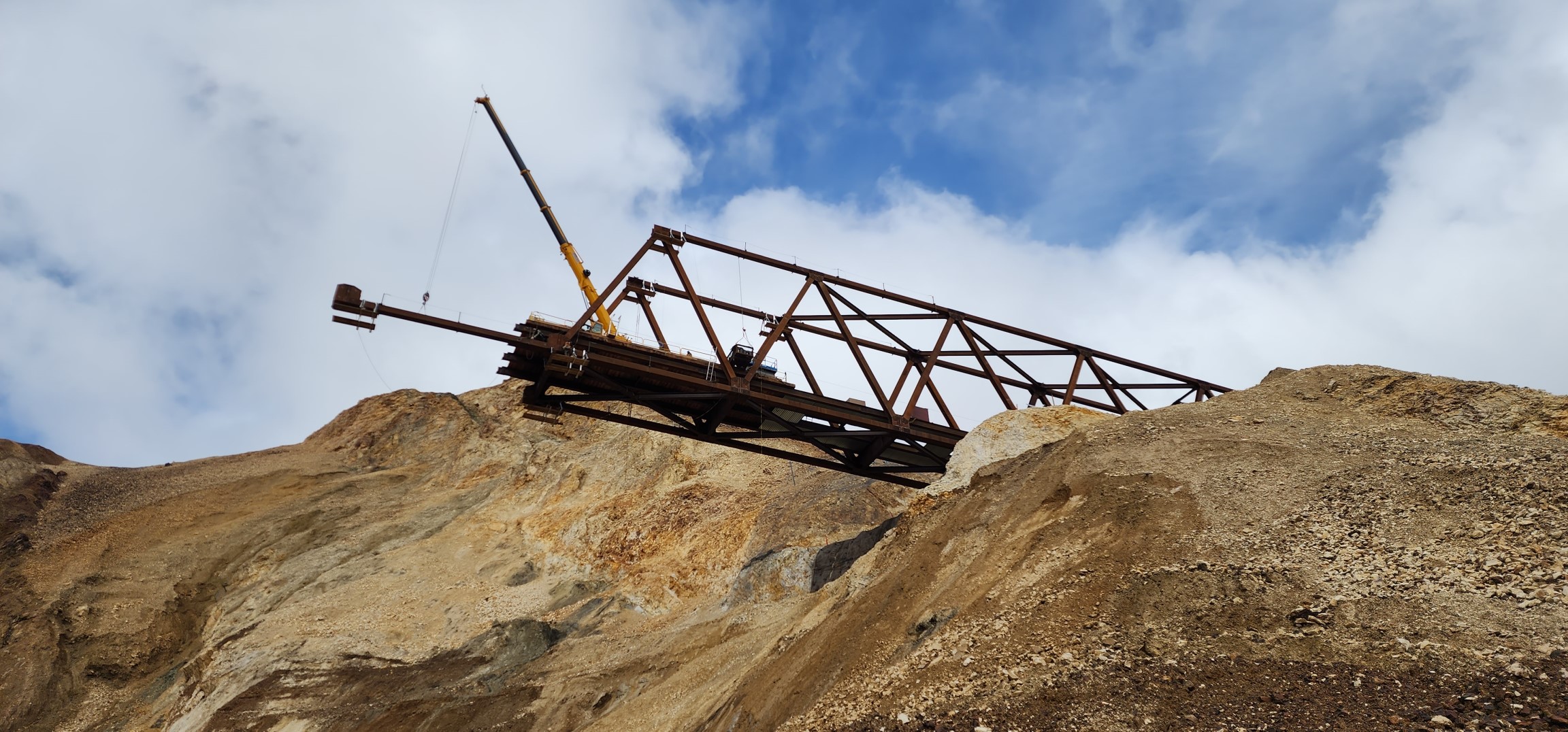 2025-06-02 hanging L2-L1 bottom chord FHWA The partial bridge that has been constructed extends out over the landslide and a steep, rocky mountainside. A crane places a metal beam at the end of the bridge.
