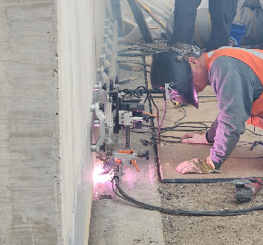 2025 09 17 Rail welding backwall proHNS Sparks fly as a welder wearing a full face helmet kneels next to the back wall of the bridge abutment.