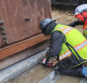2025 09 17 mag particle testing bering welds proHNS Two works wearing safety vests and helmets kneel next to large metal beams.