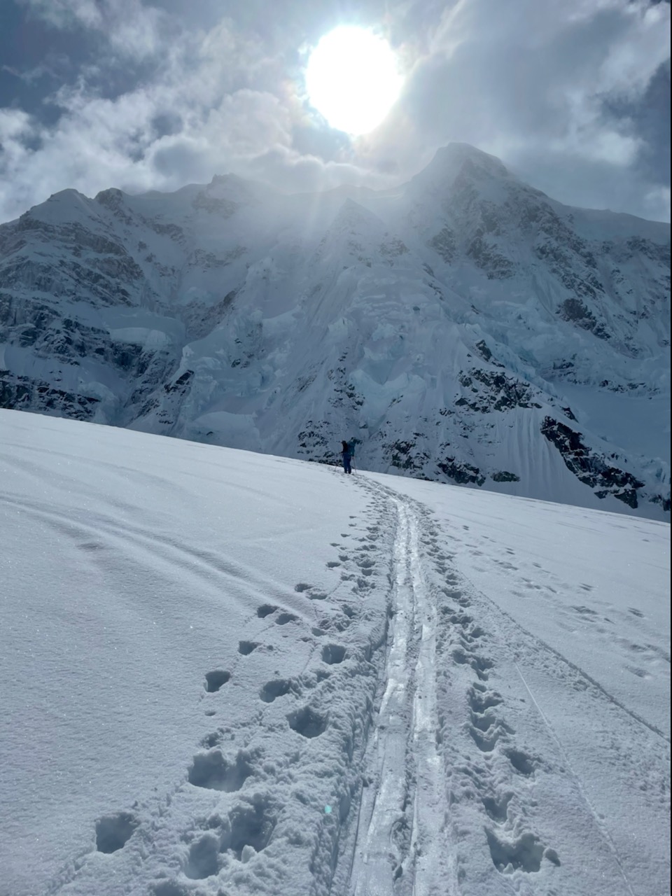 A woman skis uphill, in the background a large snowy peak with a bright sun overhead