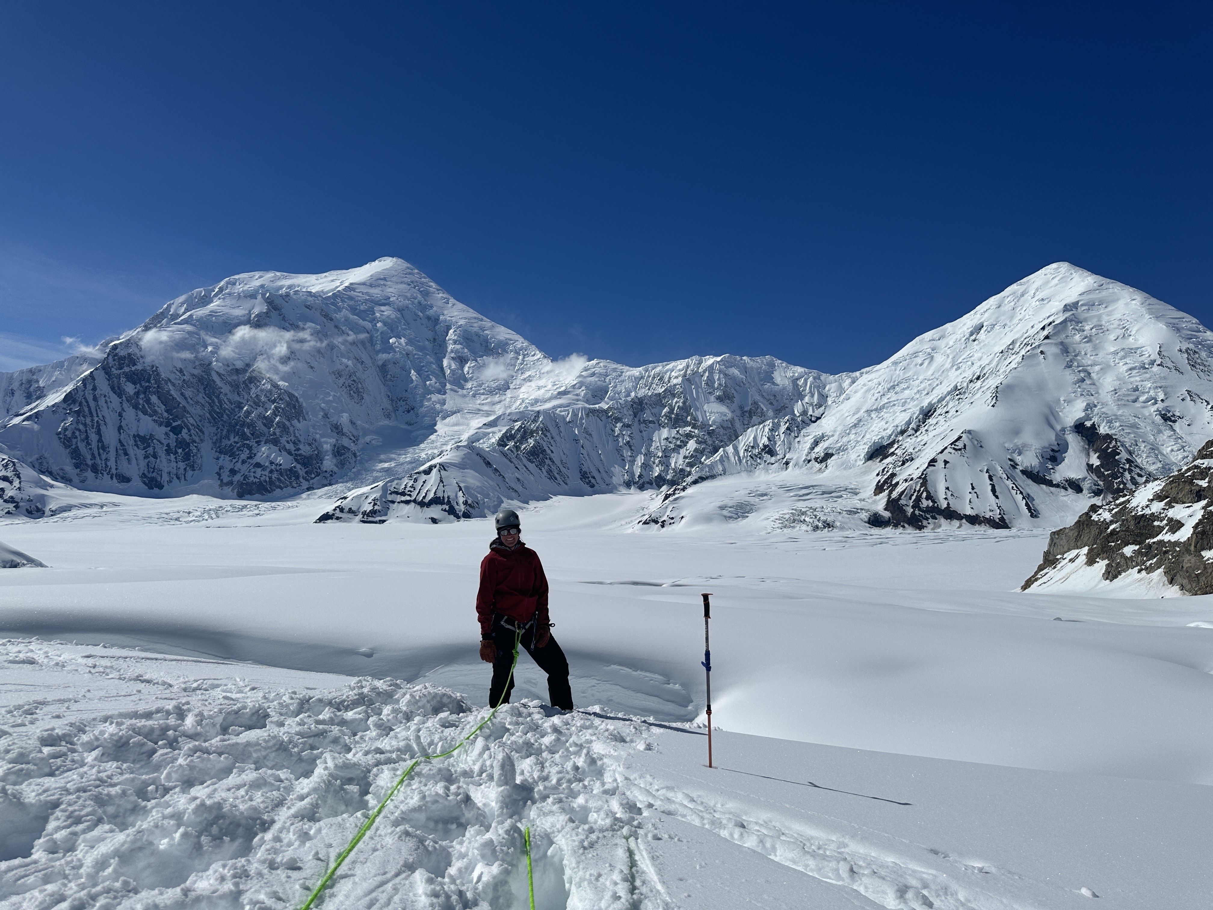 A climber stands at the edge of a glacier crevasse wearing a harness and helmet