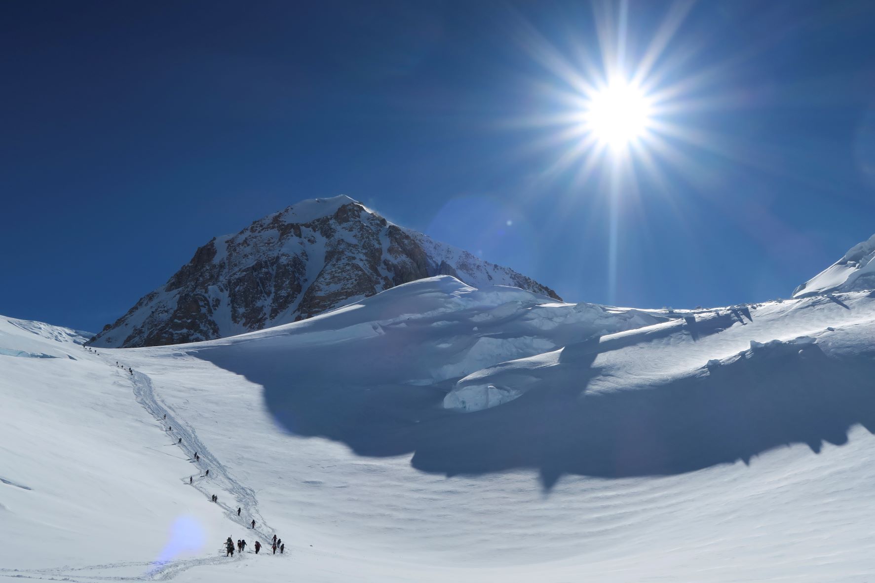 A stream of climbers ascends a hill under a bright sun