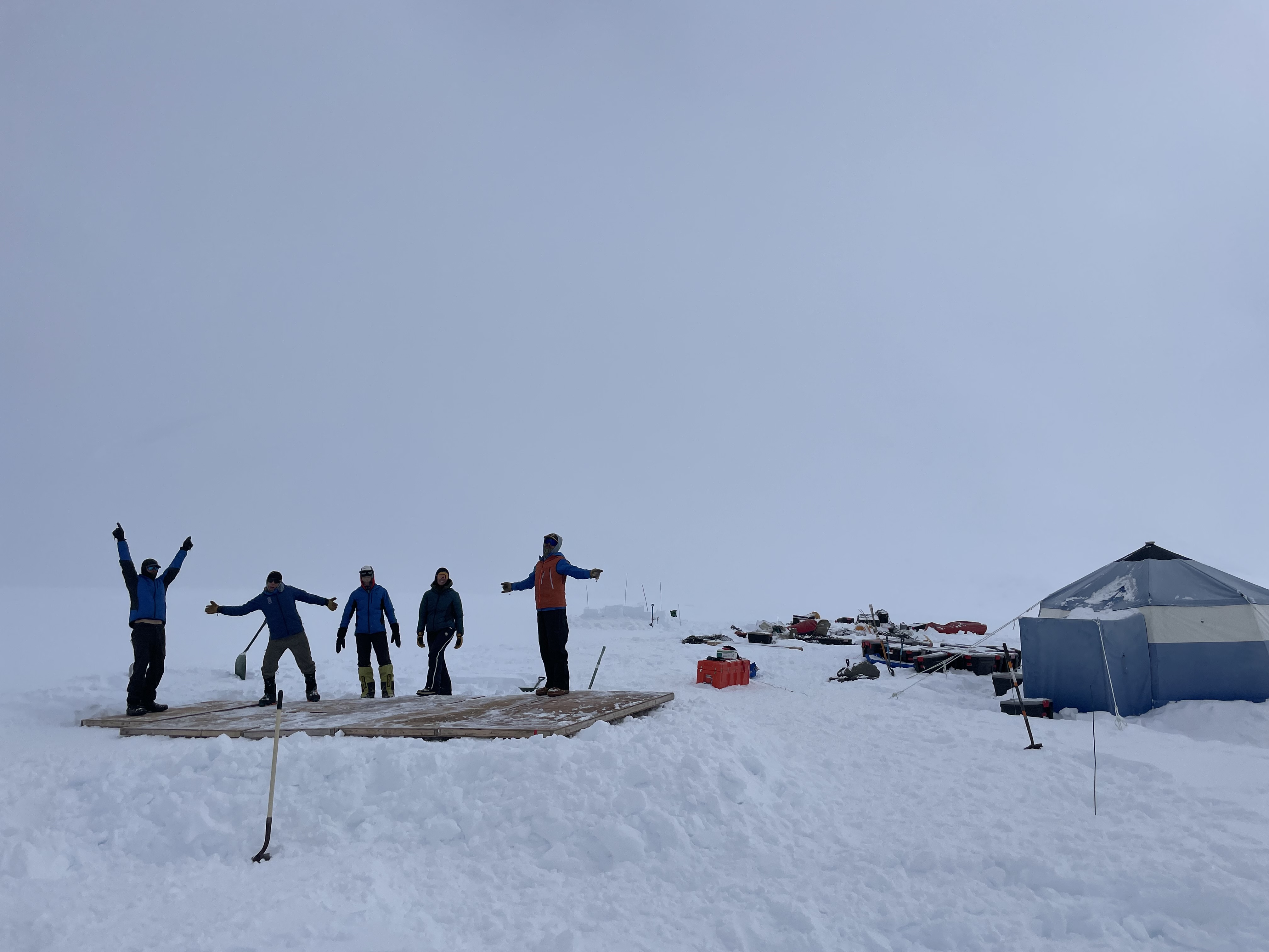 The silhouettes of five climbers stand on a wooden platform in a cloudy whiteout