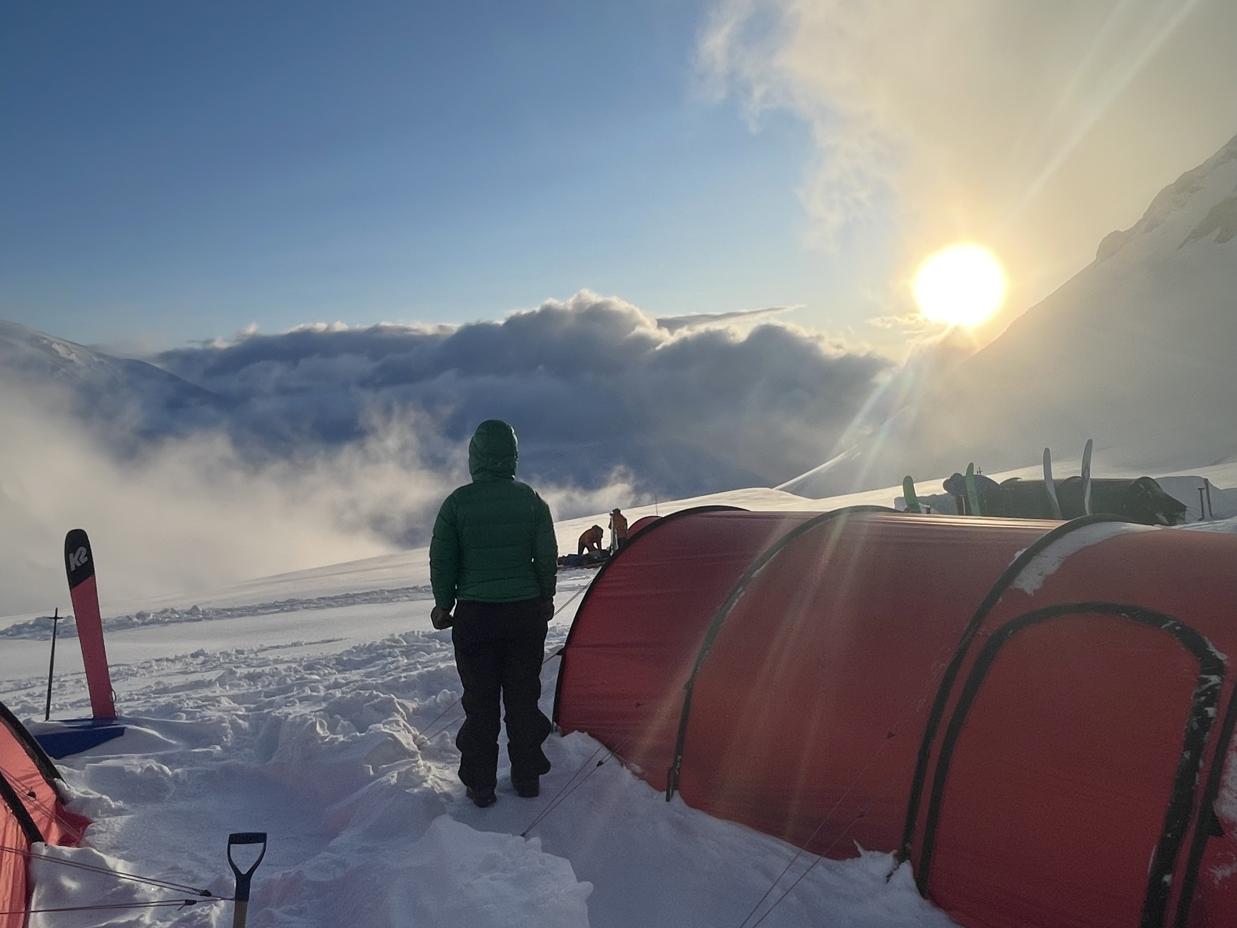 The silhouhette of a hooded woman standing next to a low red tent looking out at the sun about to set behind a mountain ridge