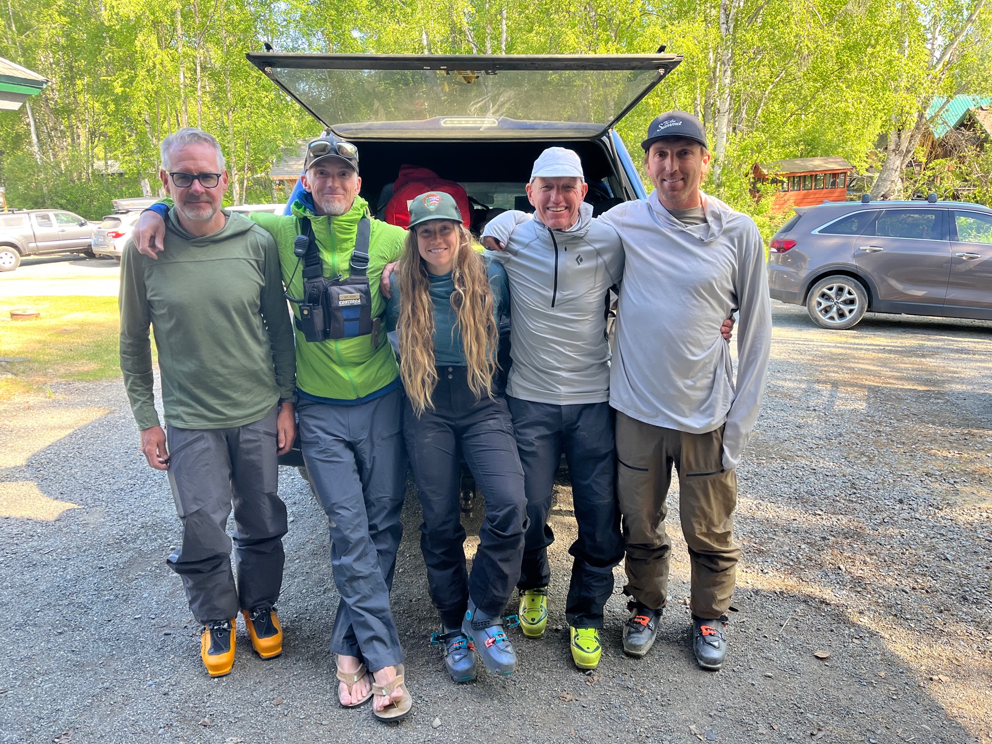 Five smiling climbers sit on the back of a pickup truck filled with duffel bags