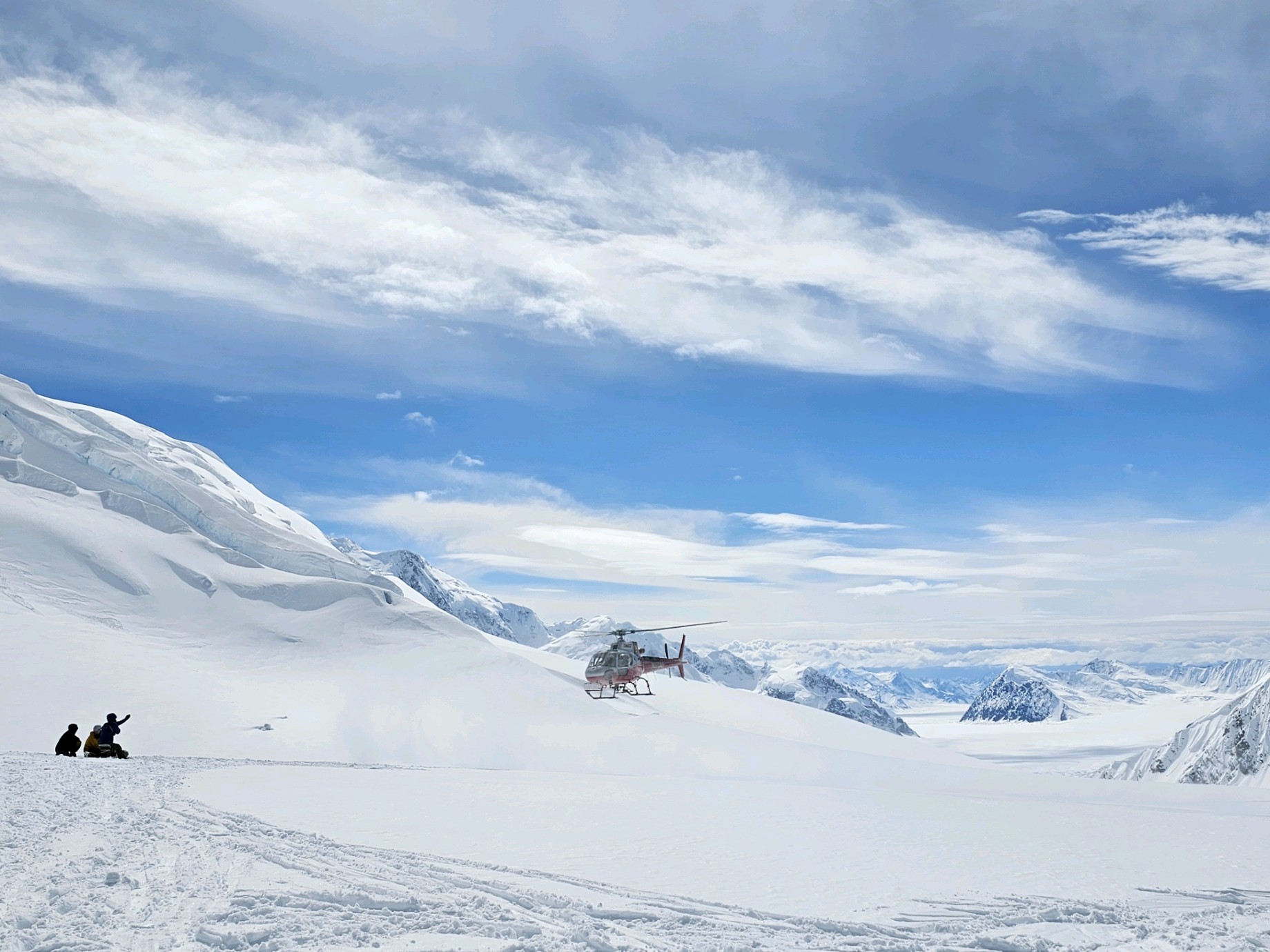 A helicopter flies onto a glacier where three climbers are kneeling, one with their arm raised