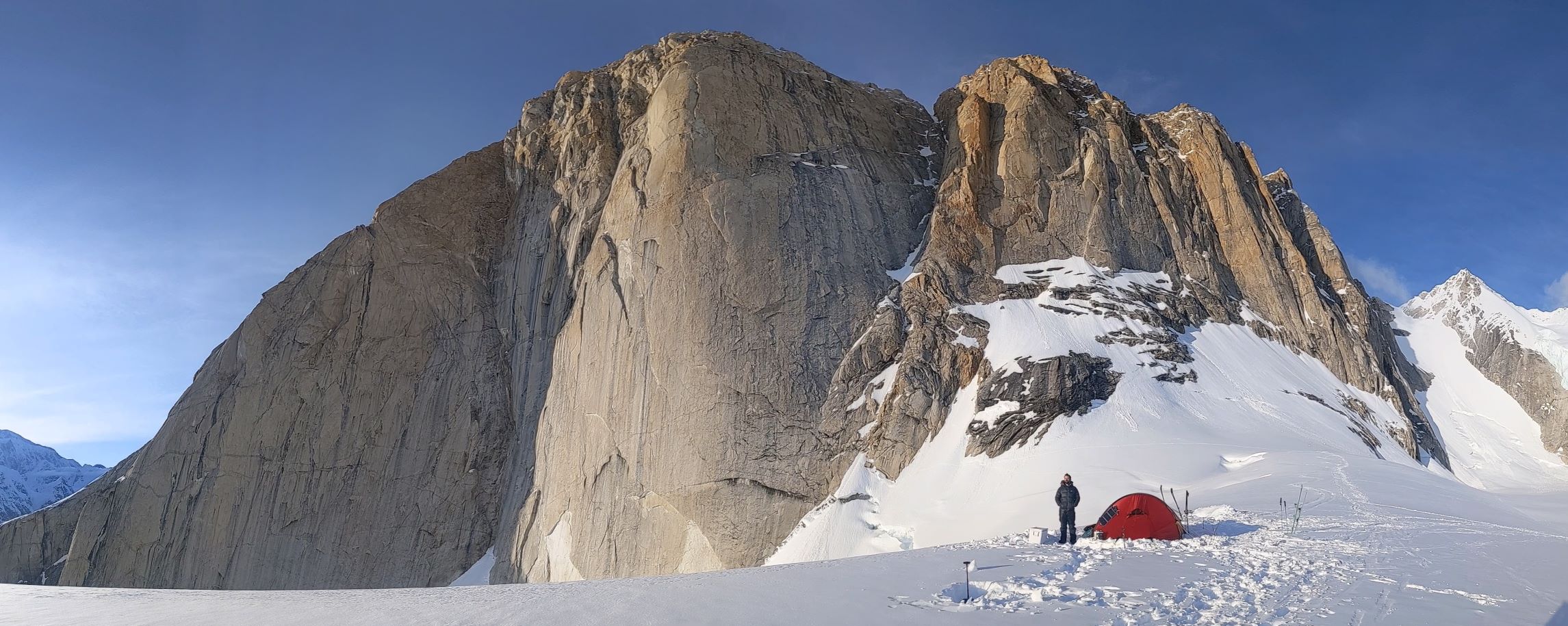 Climber stands next to his tent at the base of a dramatic rocky peak