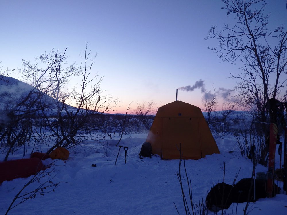 a tent set up in a snowy forest, with a small stove pipe issuing smoke from it