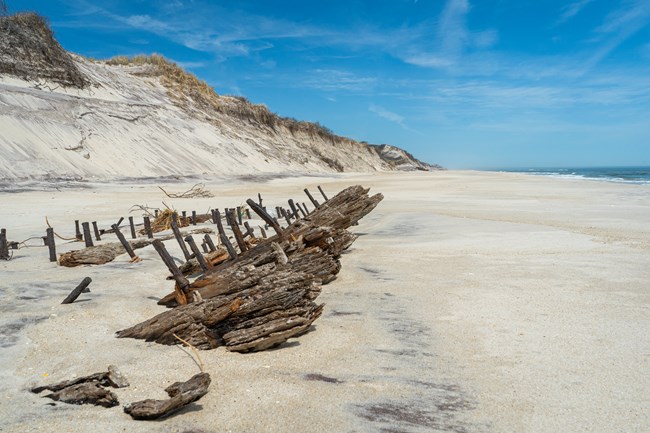 Wooden beams and large spikes protude from a sandy beach with sand dunes in the background.