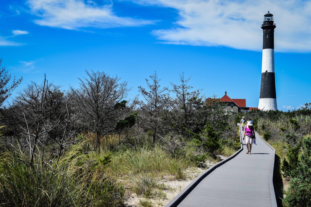 People walk on a boardwalk with a lighthouse tower in the background.