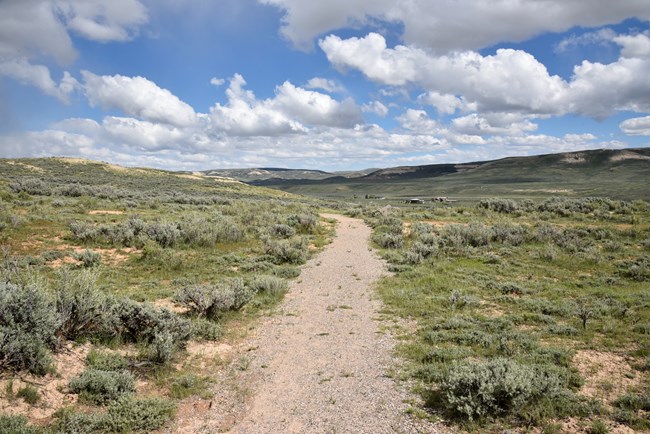 A dirt path through sagebrush leads straight away. There are small buildings visible in the distance on the right. The sky is bright blue with puffy white clouds.