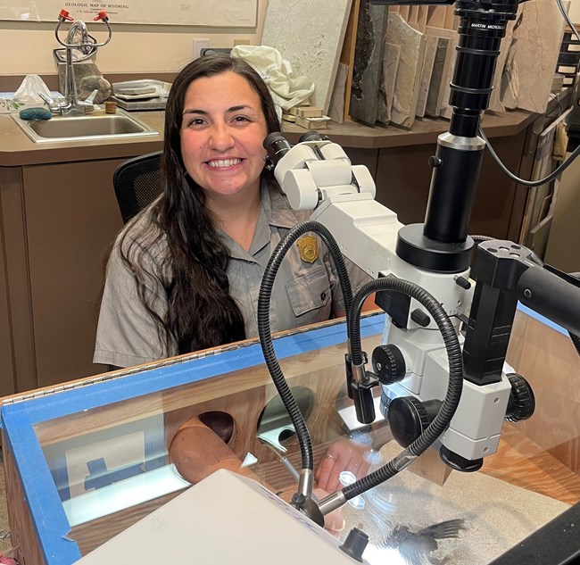 A park ranger smiles as she sits at a microscope with a fossil underneath.