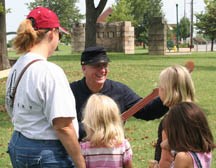 Three children and a women stand in front of a male volunteer in a union soldier uniform plays a banjo.