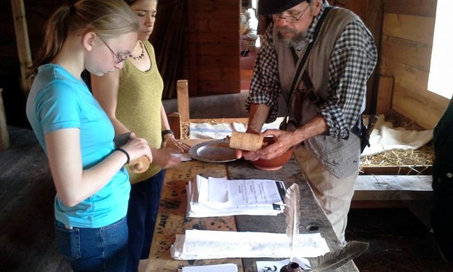 A girl and a soldier stand across a wooden table looking at a wooden cylinder.