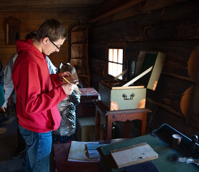 A young man stands over a table. He holds a object in his hands and studies it intently.