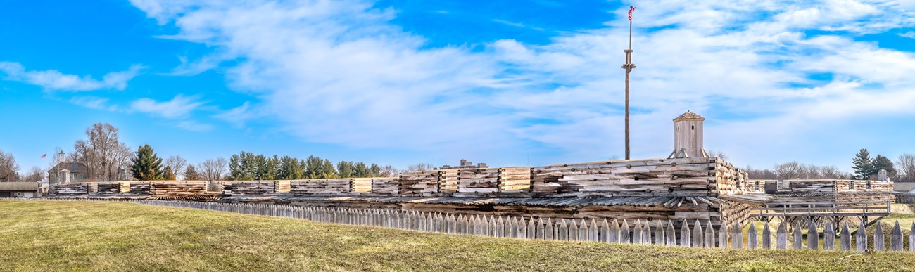 Stretched in front of you horizontally, pickets stick out behind a grass wall, and a large wooden, log cabin-style wall rises.