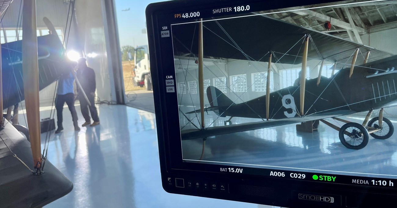 In the foreground, a screen shows the scene a camera is capturing, of an aircraft sitting inside Pearson Air Musuem's Historic Hangar. In the background, a less focused part of the image shows the front of the aircraft and pilot standing at the front.