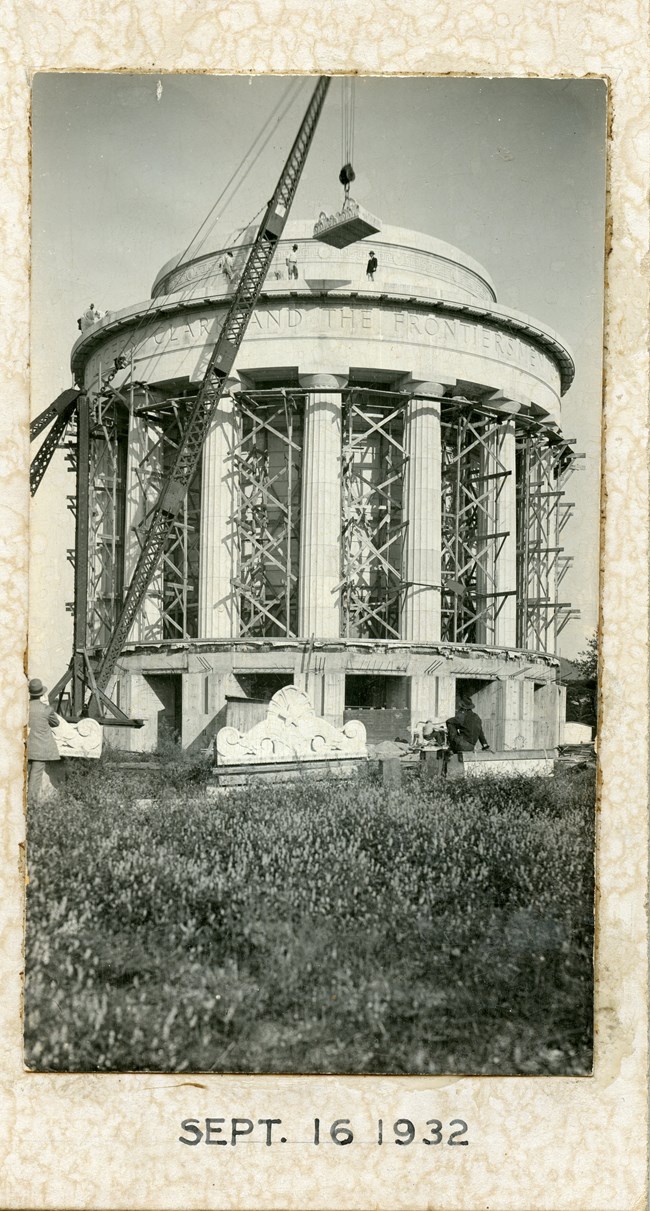 A decorative parapet section is lifted into place on the roof of the memorial, two construction workers stand ready to put it in place.