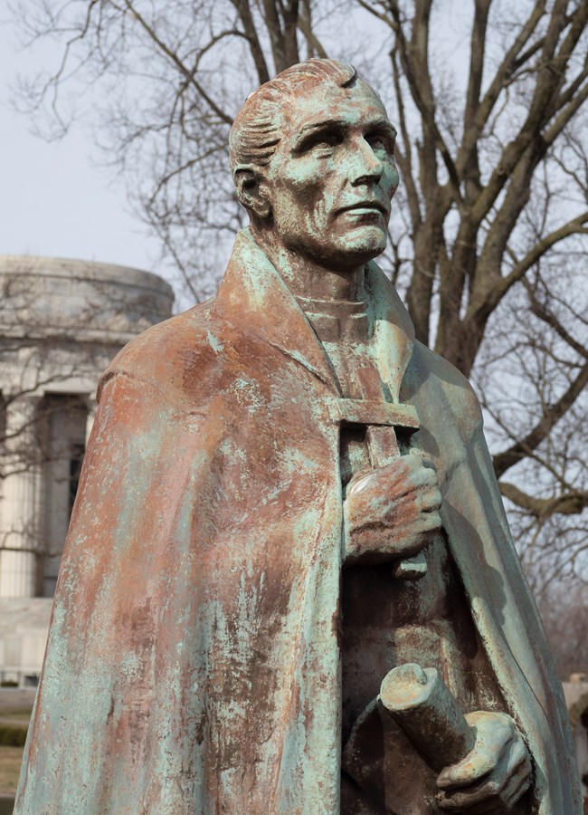 A close up image of the face of a bronze statue, the face is narrow with prominent cheeks and nose, short hair and holding a cross.