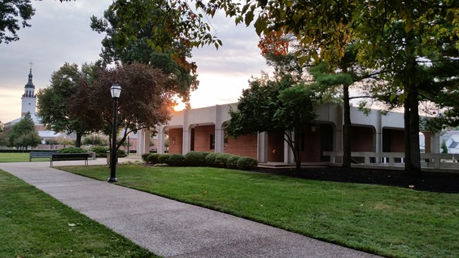 A small, square, brick and concrete building surrounded by grass, trees and sidewalks