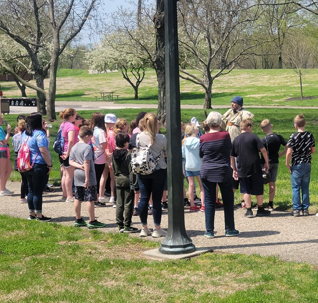 A park ranger dressed in 18th century style clothing stands in front of a group of children and adults whose backs are to the camera