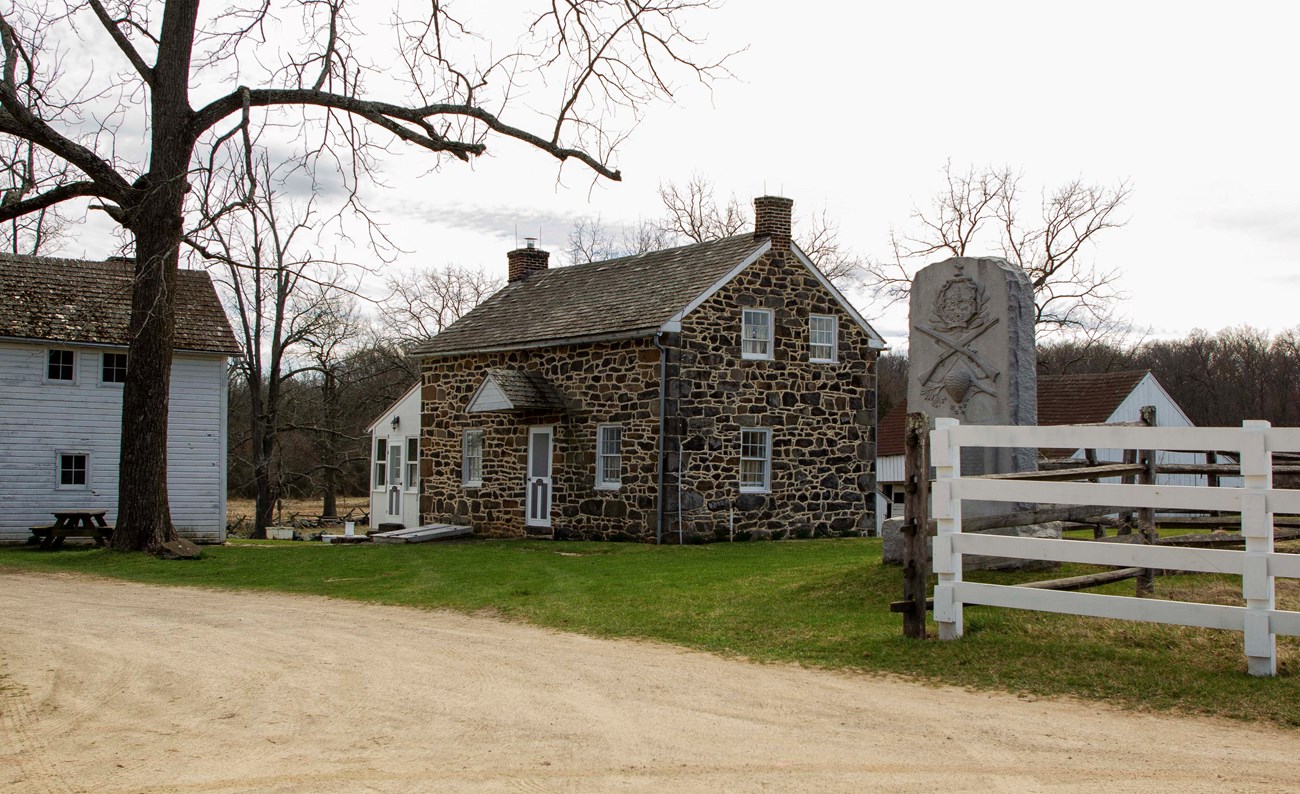 A dark stone, two-story house sits a short distance back from a light tan gravel road. There are other white sided buildings to the left and right of the house. There is a large bare tree on the left and a white board fence on the right.