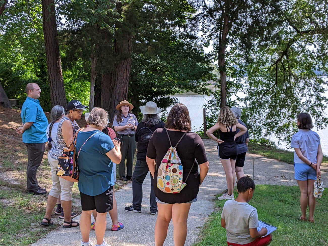 A ranger stands in front of a crowd of people during a guided tour on a trail by a large creek.