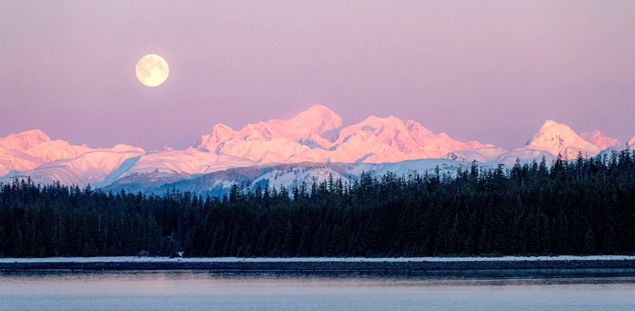 Mount Fairweather A bright full moon sits to the left above Mt Fairweather, snow colored pink by alpenglow. Bartlett Cove in foreground.