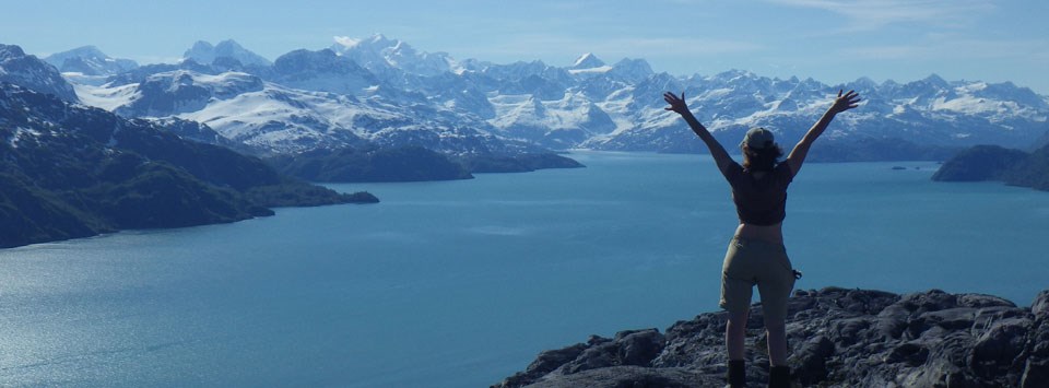 Person stands on a rock overlooking a large blue bay of water with huge snowcapped mountains rising from the water beyond them.