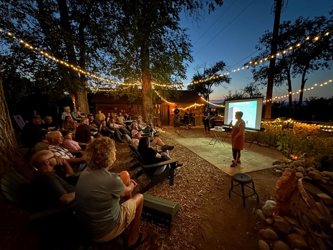 A person holds a microphone at night on a small patio in front of a crowd. Behind her is a screen with the text "Astronomy on Tap"