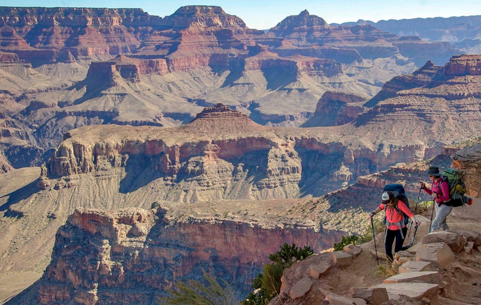 two backpackers with trekking poles are descending a unpaved trail into a desert landscape of colorful peaks, ridgelines and cliffs.