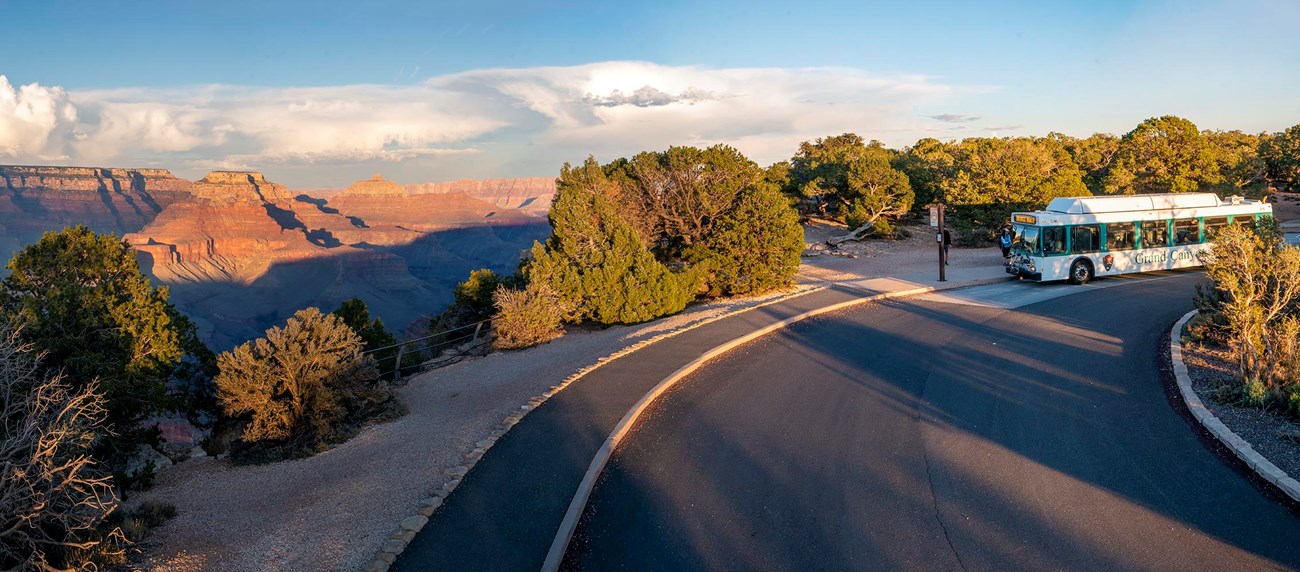 a white and green bus at a marked stop on the edge of a vast canyon landscape, just before sunset.