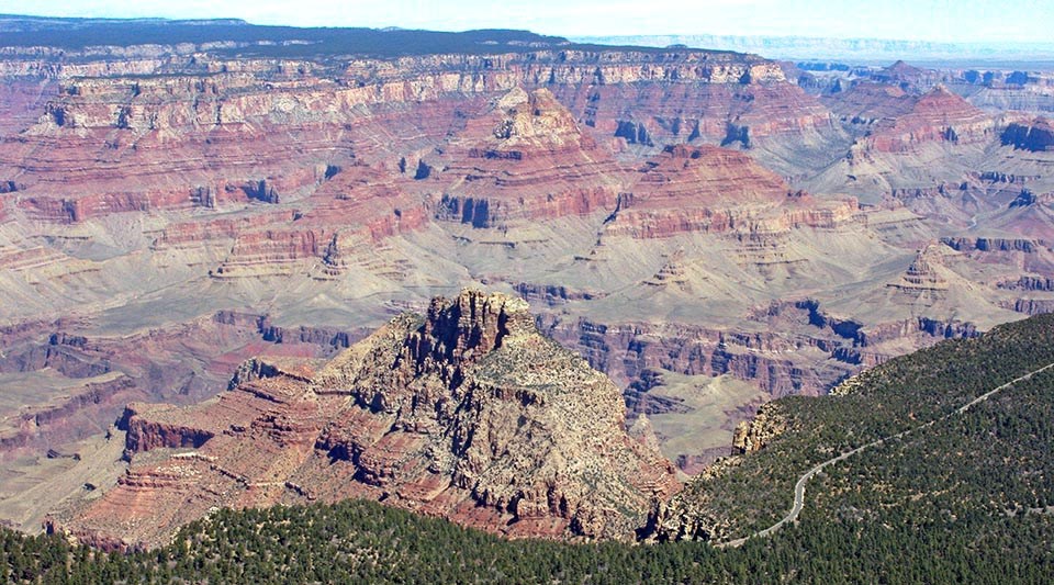 aerial view above a paved road traveling through a forested plateau on the edge of a mile deep canyon filled with colorful peaks and cliffs.