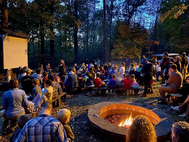 Drummers play for a crowd of people seated on benches near a burning campfire.