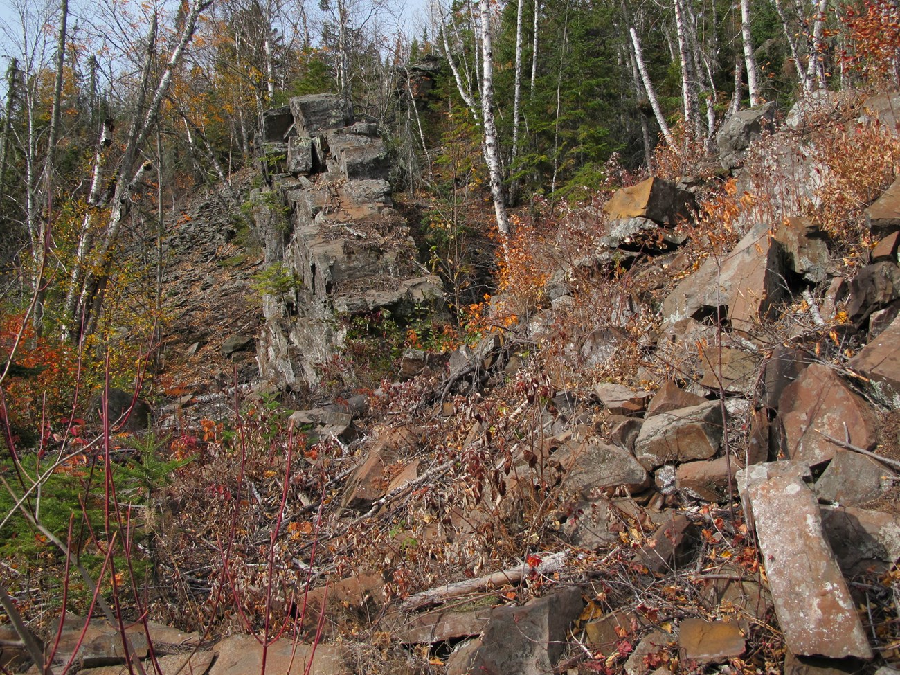 A pile of reddish chunks of rock on a hill in front of a natural gray-green rock walk.