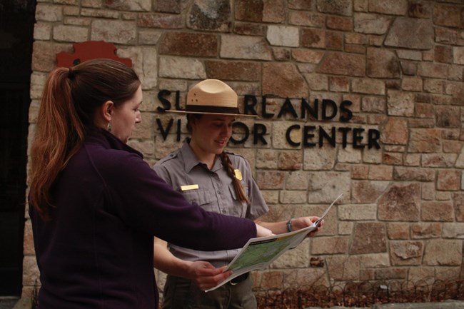 A visitor and a park ranger looking at the park newspaper in front of a visitor center.
