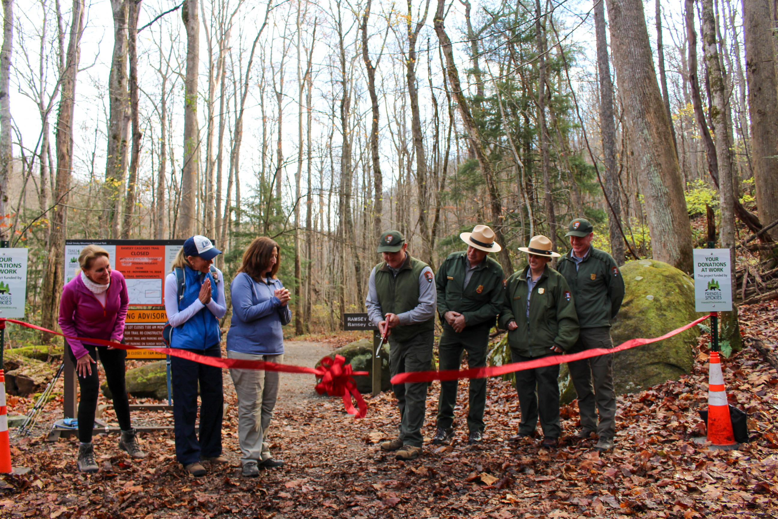 Ramsey Cascades Ribbon Cutting A red ribbon with a bow being cut and falling in front a group of park rangers and Friends of the Smokies staff. Fallen leaves, trees, and a moss-covered rock are in the background.