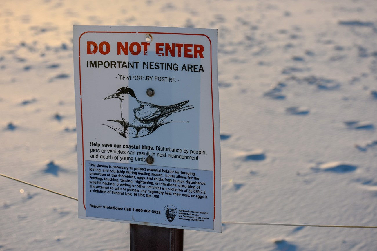 A sign with closure information stands on a post on a white sand beach.