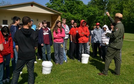 A ranger in a tan flat hat stands in front of kids while holding an object and speaking to them.