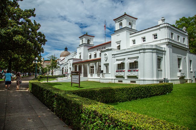 The Ozark Bathhouse on a sunny day with green grass and neatly trimmed hedges along the sidewalk in front of the building