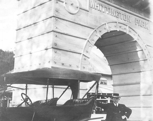 A driver dressed in suit, jacket, vest, and striped top hat poses with his 1920s style black convertible carriage car in front of a tall white stone archway that says "whittington park".