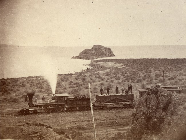 This historic image captures a locomotive and work crew near Monument Point, with the Great Salt Lake in the background—part of the final push to complete the transcontinental railroad in 1869. Steam locomotive passes near shoreline of Great Salt Lake with workers standing on flatcars