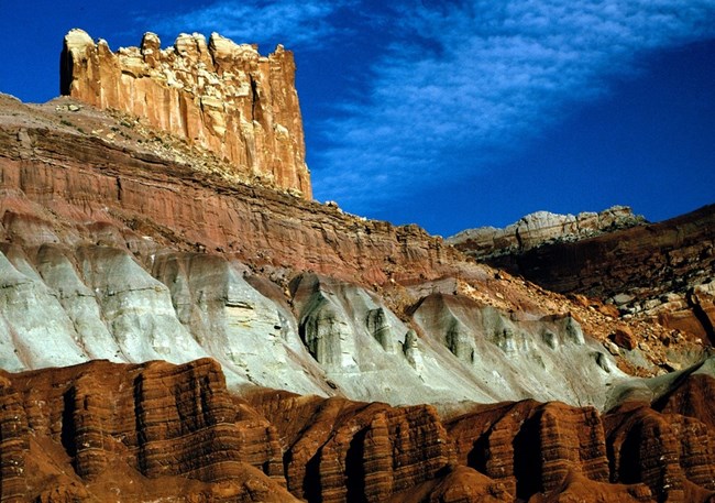 Capitol Reef’s iconic Castle formation showcases geologic layers shaped by millions of years of erosion. The Castle rises above striped red and gray rock layers under a deep blue sky.