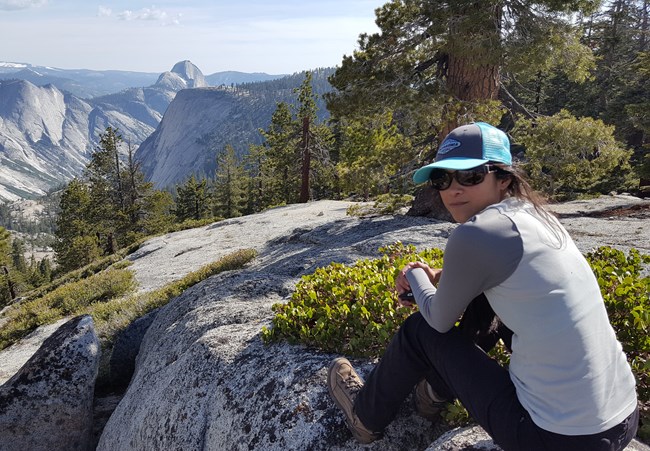 Woman sitting on granite outcrop, looking back at camera. Views of large granite domes in background.