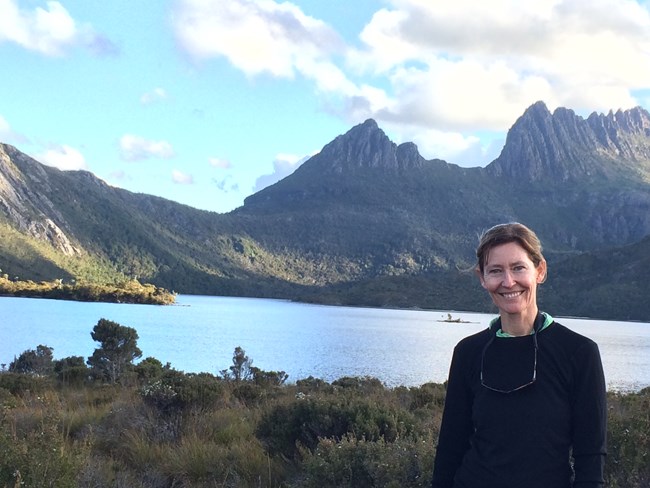 Woman stands in front of a scenic lake with rugged mountains in the background.