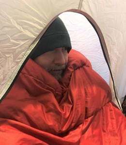 A man wearing a beanie is in a tent with a heavy orange blanket covering him.