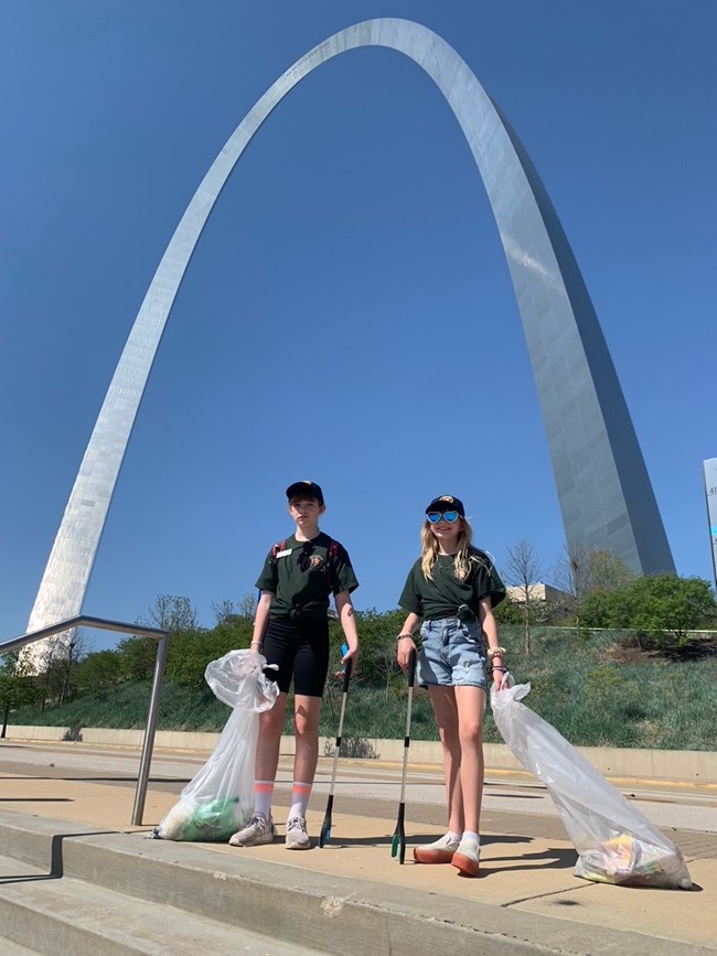 Two girls in green Volunteer shirts hold trash bags and litter picker sticks. The Gateway Arch is in the background.